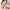 A woman in a white robe applies a pink cream from a red jar to her cheek. The jar has Korean text on the label and its red lid lies beside it on a marble countertop in a bright bathroom setting.